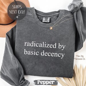 May include: A dark gray sweatshirt with the text "radicalized by basic decency" in white. The sweatshirt has a crew neck and long sleeves. A gold pendant necklace is draped on the shirt. The word "Pepper" is at the bottom.