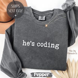 Może przedstawiać: Szara bluza w kolorze pieprzu z białym napisem "he's coding". Bluza ma okrągły dekolt i długie rękawy. Złoty naszyjnik z wisiorkiem jest zarzucony na koszulę. Widoczne są słowa "The Ivy Threads Co.".