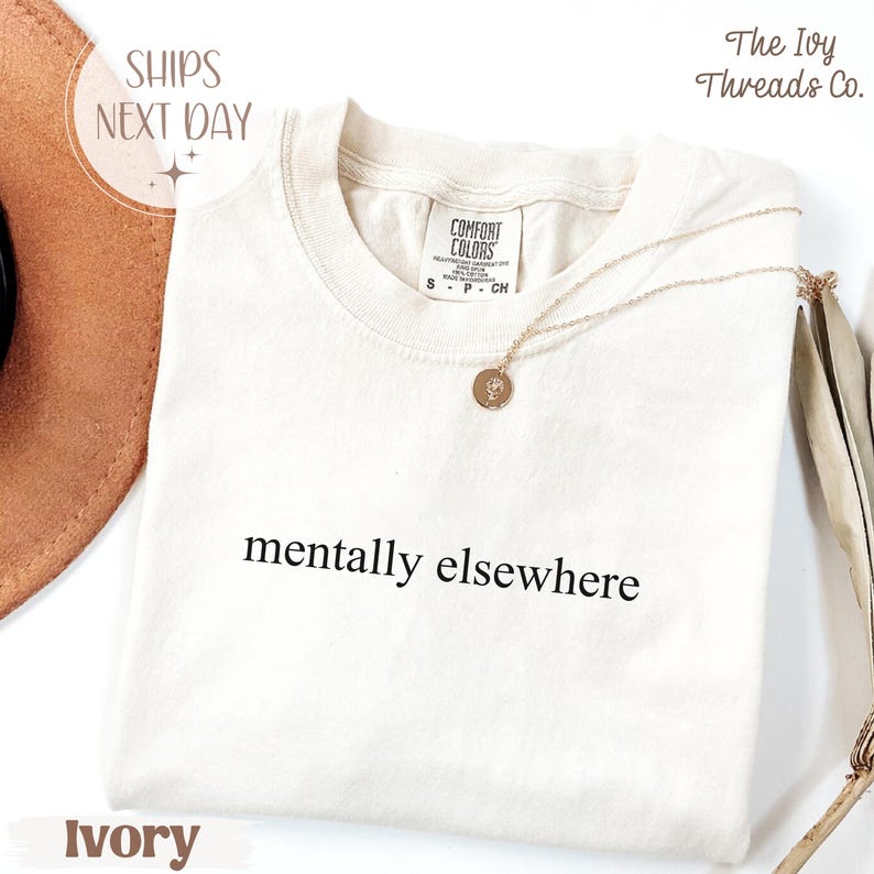May include: Ivory-colored t-shirt with the phrase "mentally elsewhere" in black. A gold necklace with a pendant rests on the shirt. The shirt has a Comfort Colors tag. The words "Ships Next Day" are in the upper left corner.