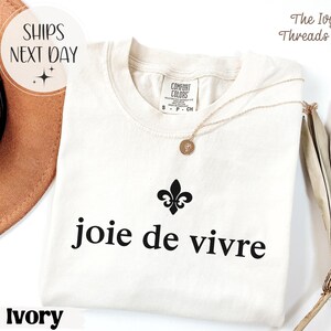 Puede incluir: Camiseta de color marfil con la frase "joie de vivre" en negro, una flor de lis negra y un collar dorado. La camiseta est&aacute; hecha de un material suave. El texto "SHIPS NEXT DAY" est&aacute; en un c&iacute;rculo.