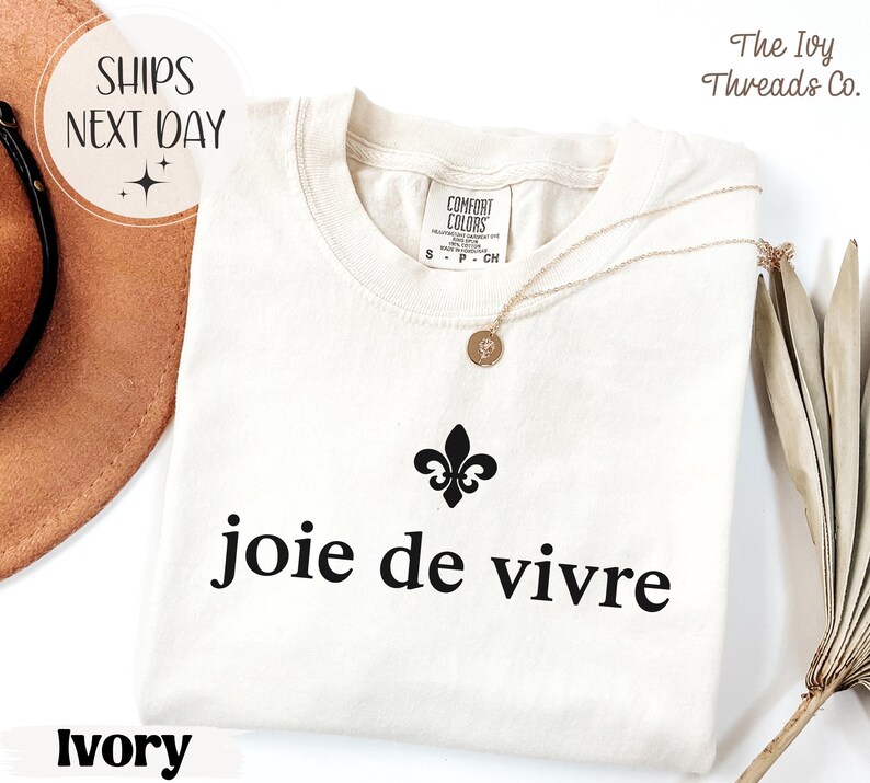 Puede incluir: Camiseta de color marfil con la frase "joie de vivre" en negro, una flor de lis negra y un collar dorado. La camiseta est&aacute; hecha de un material suave. El texto "SHIPS NEXT DAY" est&aacute; en un c&iacute;rculo.