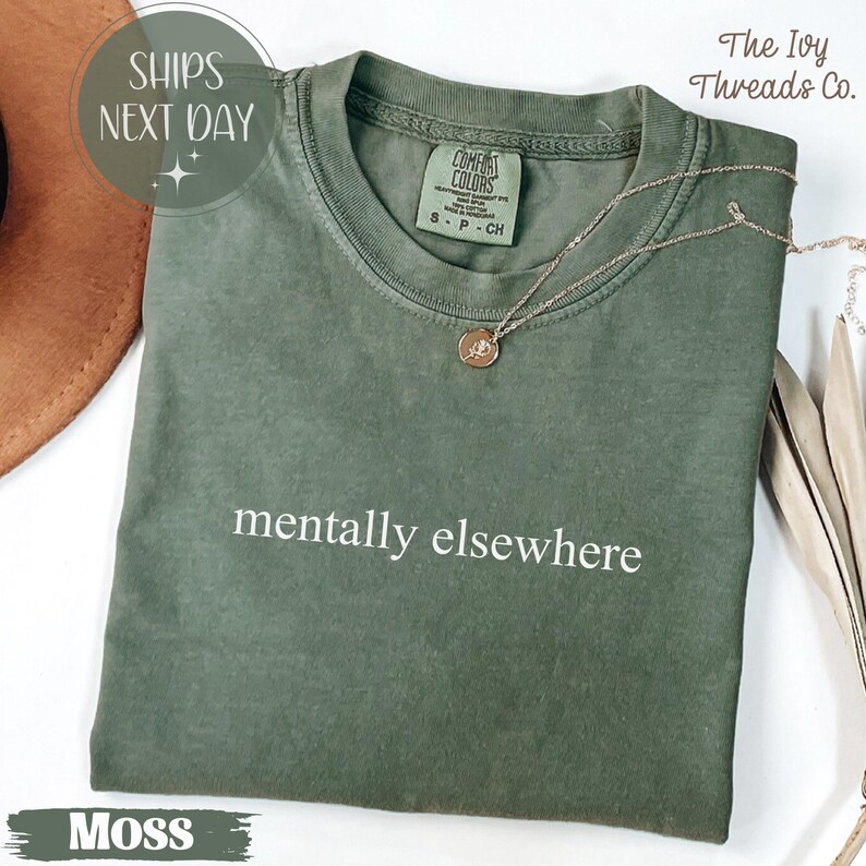 May include: A moss green t-shirt with the phrase "mentally elsewhere" printed in white. A gold necklace with a pendant rests on the shirt. The shirt is from The Ivy Threads Co.
