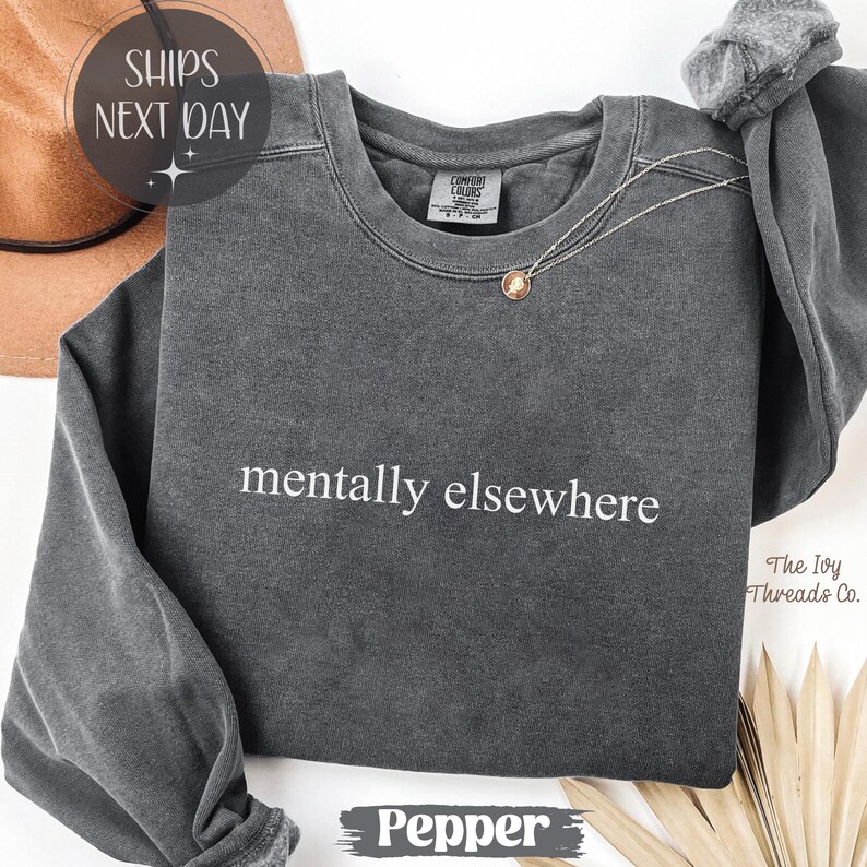 May include: A dark gray sweatshirt with the phrase "mentally elsewhere" in white lettering. A gold pendant necklace is draped on the front. The sweatshirt is labeled "Pepper" at the bottom.