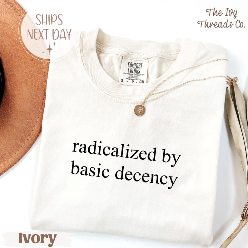 May include: Ivory-colored t-shirt with the text "radicalized by basic decency" in black. A gold necklace with a pendant rests on the shirt. The shirt has a Comfort Colors tag and the words "SHIPS NEXT DAY" are visible.