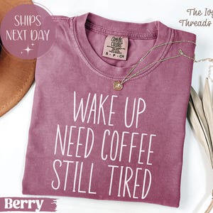 May include: Berry-colored t-shirt with the text "WAKE UP NEED COFFEE STILL TIRED" in white. A gold necklace with a pendant rests on the shirt. The shirt has a Comfort Colors tag.