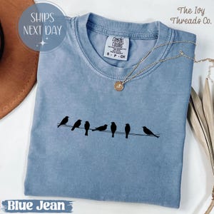 Könnte beinhalten: Ein blaues Comfort Colors T-Shirt mit einem schwarzen Aufdruck von Vögeln auf einem Draht. Eine goldene Halskette mit Anhänger liegt auf dem Shirt. Der Text "Blue Jean" befindet sich unten.