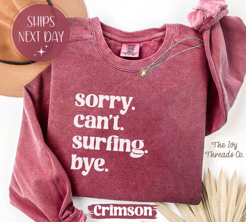 May include: Crimson-colored sweatshirt with the text "sorry. can't. surfing. bye." in white. A gold necklace and a brown hat are also visible. The Ivy Threads Co. is printed on the bottom right.