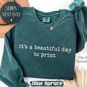Può includere: Una felpa color verde acqua con la scritta bianca "it's a beautiful day to print". Una collana d'oro è drappeggiata sul davanti. Il testo "Blue Spruce" è in basso. Marchio Comfort Colors.