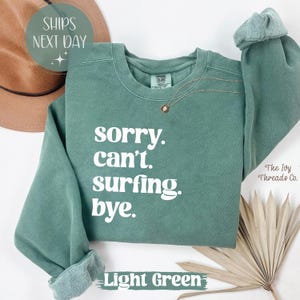 May include: Light green sweatshirt with the text "sorry. can't. surfing. bye." in white. The sweatshirt has a crew neck and long sleeves with rolled cuffs. A gold necklace with a small pendant is visible.