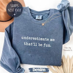 Könnte beinhalten: Denimfarbenes Sweatshirt mit dem Text "underestimate me that'll be fun." Eine goldene Halskette mit Anhänger liegt auf dem Shirt. The Ivy Threads Co. ist unten rechts aufgedruckt. Die Worte "SHIPS NEXT DAY" stehen in einem Kreis oben links.