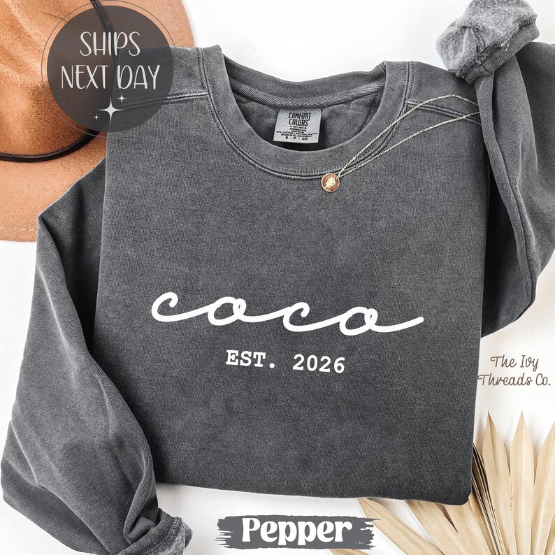 May include: A dark gray sweatshirt with the word "coco" in white script and the text "EST. 2026" below it. A gold pendant necklace is draped on the front. The sweatshirt is a Comfort Colors brand. The word "Pepper" is at the bottom.
