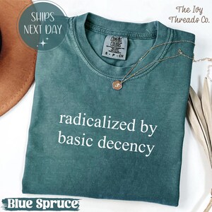 May include: A blue spruce Comfort Colors t-shirt with the phrase "radicalized by basic decency" in white. A gold necklace with a pendant rests on the shirt. The Ivy Threads Co. logo is in the upper right corner.