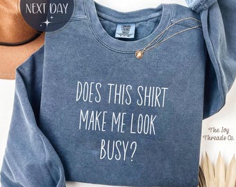May include: Denim blue sweatshirt with the text "DOES THIS SHIRT MAKE ME LOOK BUSY?" in white. A gold necklace with a pendant is draped on the shirt. The Ivy Threads Co. logo is visible.