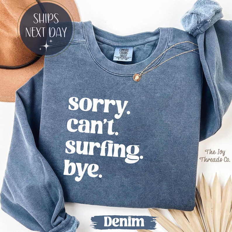 May include: A denim blue sweatshirt with the text "sorry. can't. surfing. bye." in white. The sweatshirt has a crew neck and long sleeves. A gold necklace with a pendant is draped on the shirt.