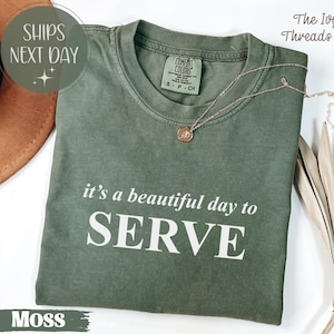 Könnte beinhalten: Ein moosgrünes T-Shirt mit dem weißen Text "it's a beautiful day to SERVE". Eine goldene Halskette mit Anhänger liegt auf dem Shirt. Das Shirt ist von The Ivy Threads Co.