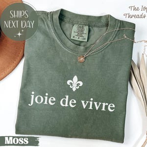 Puede incluir: Camiseta verde musgo con la frase "joie de vivre" en blanco y un s&iacute;mbolo de flor de lis. Un collar dorado descansa sobre la camiseta. La etiqueta dice "Comfort Colors" y "S-P-CH". El texto "SHIPS NEXT DAY" tambi&eacute;n es visible.
