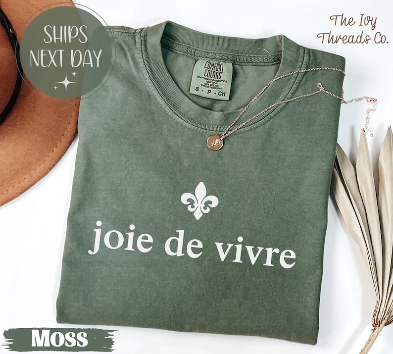 Puede incluir: Camiseta verde musgo con la frase "joie de vivre" en blanco y un s&iacute;mbolo de flor de lis. Un collar dorado descansa sobre la camiseta. La etiqueta dice "Comfort Colors" y "S-P-CH". El texto "SHIPS NEXT DAY" tambi&eacute;n es visible.