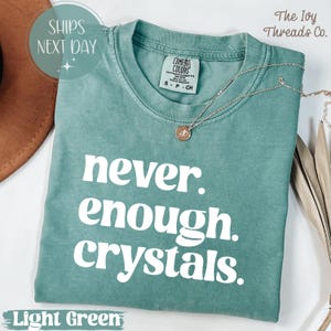 May include: Light green t-shirt with the text "never enough crystals" in white. The shirt has a crew neck and short sleeves. A silver necklace with a pendant is draped across the shirt. The shirt is folded on a white surface.