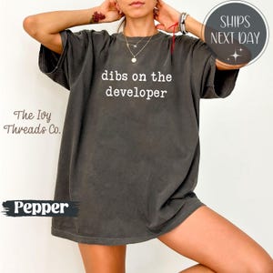 May include: A dark grey t-shirt with the phrase "dibs on the developer" printed in white. The Ivy Threads Co. logo is on the left. A circular graphic with "SHIPS NEXT DAY" is on the upper right.