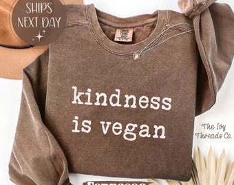 May include: Espresso-coloured sweatshirt with the text "kindness is vegan" in white. The long-sleeved top has a crew neck and a Comfort Colors tag. A gold necklace is draped on the front. The Ivy Threads Co. logo is visible.
