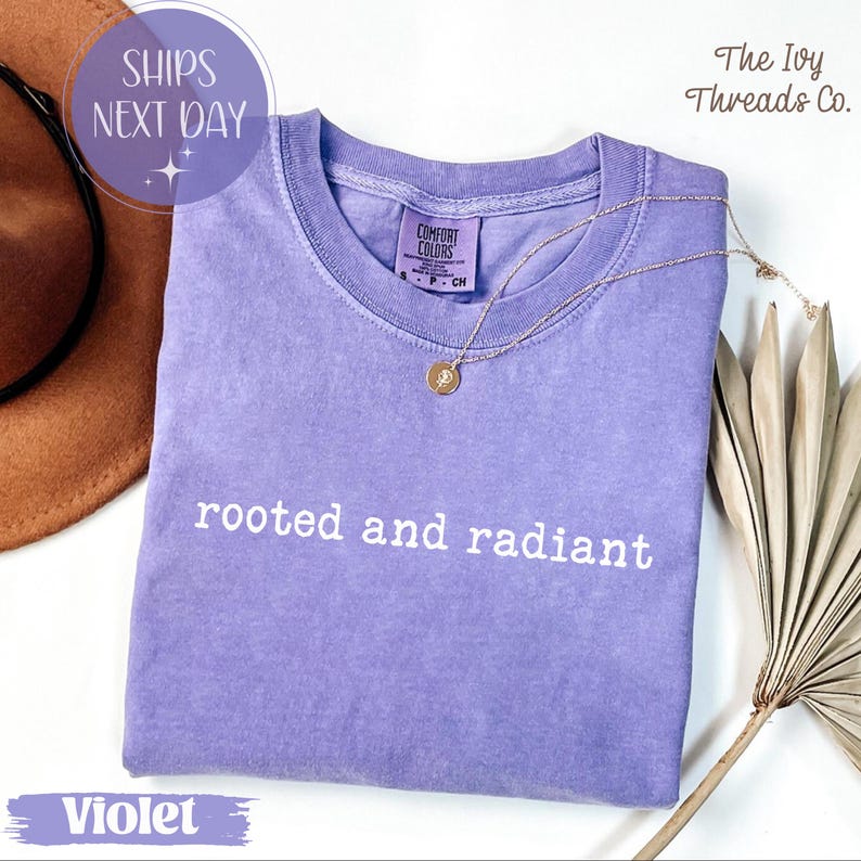 May include: A violet Comfort Colors t-shirt with the words "rooted and radiant" in white. A gold necklace with a pendant rests on the shirt. The Ivy Threads Co. logo is in the upper right corner.