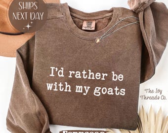 Peut inclure: Sweat-shirt couleur expresso avec l'inscription blanche "I'd rather be with my goats". Le haut à manches longues a un col rond et une coupe décontractée. Un collier doré est drapé sur le sweat-shirt.