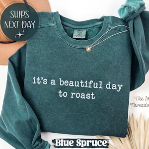 May include: A teal blue sweatshirt with the phrase "it's a beautiful day to roast" in white lettering. The sweatshirt has a crew neck and long sleeves. A gold necklace is draped on the front. The words "Blue Spruce" are at the bottom.