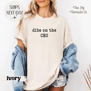 May include: Ivory-colored t-shirt with the text "dibs on the CEO" in black. The shirt is a relaxed fit and has rolled-up sleeves. A denim jacket is draped over the shoulders. The Ivy Threads Co. logo is visible.