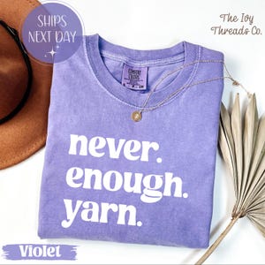 May include: A violet Comfort Colors t-shirt with the text "never. enough. yarn." in white. A gold necklace with a pendant rests on the shirt. The shirt is part of a collection from The Ivy Threads Co.