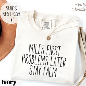 以下が含まれることがあります： 「MILES FIRST PROBLEMS LATER STAY CALM」の黒い文字がプリントされたアイボリー色のTシャツ。丸首で半袖です。金色のネックレスがTシャツの上にかけられています。The Ivy Threads Co.のロゴが右上隅にあります。
