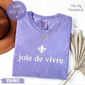 Puede incluir: Una camiseta violeta Comfort Colors con la frase "joie de vivre" en blanco, un dise&ntilde;o de flor de lis y un collar dorado. La camiseta se muestra con un sombrero marr&oacute;n y hojas de palma secas.