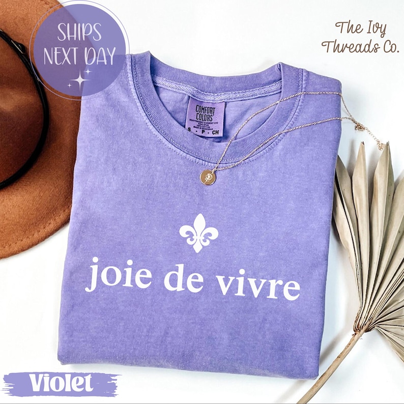 Puede incluir: Una camiseta violeta Comfort Colors con la frase "joie de vivre" en blanco, un dise&ntilde;o de flor de lis y un collar dorado. La camiseta se muestra con un sombrero marr&oacute;n y hojas de palma secas.