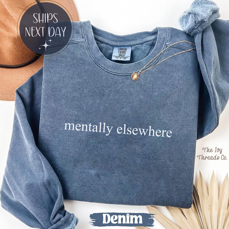 May include: Denim blue sweatshirt with the phrase "mentally elsewhere" in white. A gold necklace with a pendant rests on the shirt. The text "The Ivy Threads Co." is visible on the right side of the image.