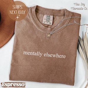 May include: Espresso-colored t-shirt with the phrase "mentally elsewhere" in white. The shirt has a crew neck and short sleeves. A silver necklace with a small pendant is draped across the shirt. The shirt is labeled "Comfort Colors."