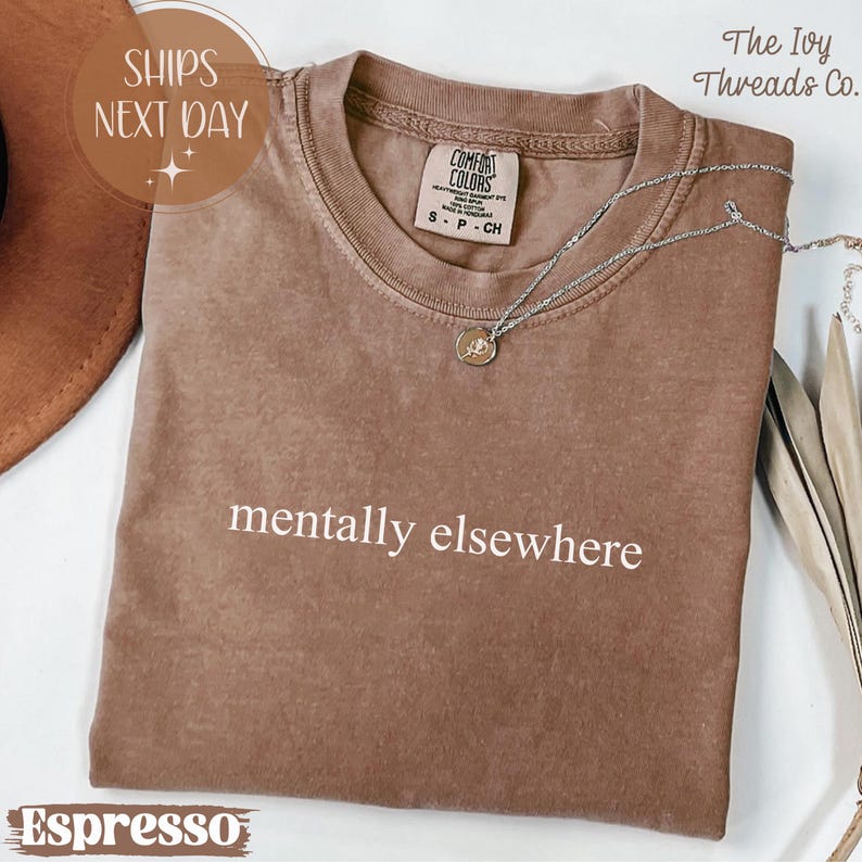 May include: Espresso-colored t-shirt with the phrase "mentally elsewhere" in white. The shirt has a crew neck and short sleeves. A silver necklace with a small pendant is draped across the shirt. The shirt is labeled "Comfort Colors."