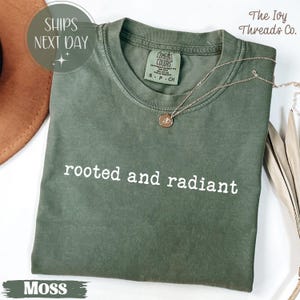 May include: A moss green t-shirt with the words "rooted and radiant" in white. The shirt has a crew neck and short sleeves. A gold necklace with a pendant rests on the shirt. The shirt is labeled "Comfort Colors" and "S-P-CH".