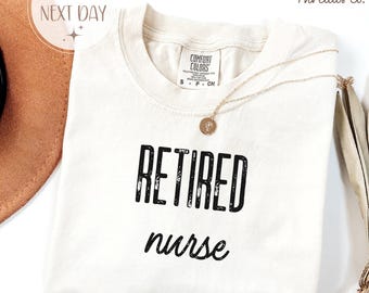 May include: Ivory-colored t-shirt with the words "RETIRED nurse" printed in black. The shirt has a crew neck and short sleeves. A gold necklace with a pendant rests on the shirt. The shirt is from The Ivy Threads Co.