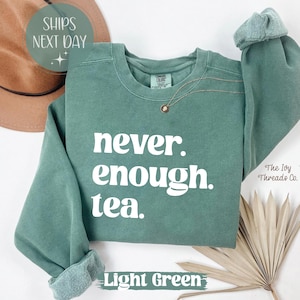 以下が含まれることがあります： 「never. enough. tea.」の白文字がプリントされた、ライトグリーンのスウェットシャツ。クルーネックと、ロールカフスの長袖。ゴールドのネックレスが見える。「Light Green」の文字が下部に表示。