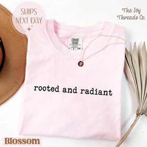 May include: A light pink t-shirt with the words "rooted and radiant" printed in black. A delicate necklace with a small pendant rests on the shirt. The shirt is labeled "Blossom" and the brand name is "The Ivy Threads Co."