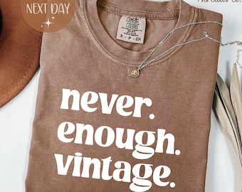 May include: Espresso-colored t-shirt with the text "never. enough. vintage." in white. The shirt has a crew neck and short sleeves. A silver necklace with a pendant is draped across the shirt. The shirt is from The Ivy Threads Co.