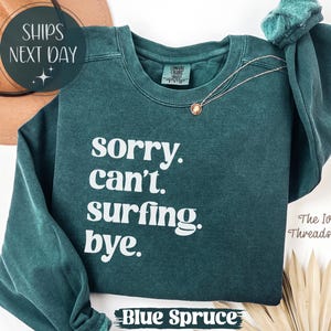 May include: A blue spruce sweatshirt with the text "sorry. can't. surfing. bye." in white. The sweatshirt has long sleeves and a crew neck. A gold necklace is draped on the front. The words "SHIPS NEXT DAY" are in a circle at the top.
