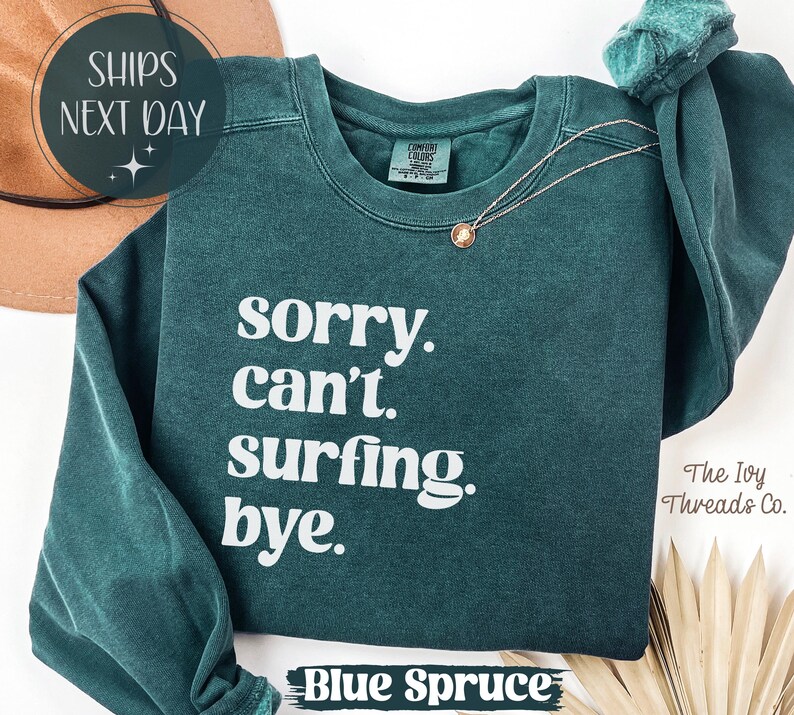 May include: A blue spruce sweatshirt with the text "sorry. can't. surfing. bye." in white. The sweatshirt has long sleeves and a crew neck. A gold necklace is draped on the front. The words "SHIPS NEXT DAY" are in a circle at the top.