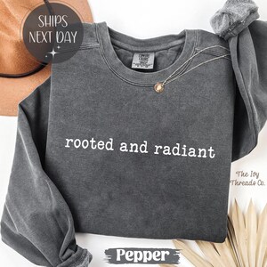 May include: A dark gray sweatshirt with the words "rooted and radiant" in white. The sweatshirt has a crew neck and long sleeves. A gold pendant necklace is draped on the front. The word "Pepper" is at the bottom.