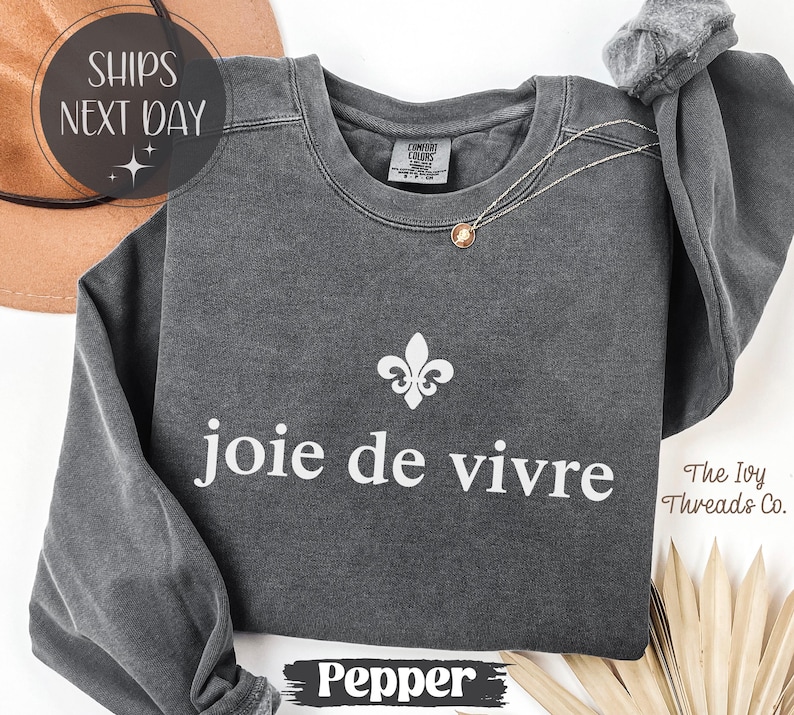 Puede incluir: Una sudadera gris oscuro con la frase "joie de vivre" en blanco y un s&iacute;mbolo de flor de lis. La sudadera tiene mangas largas y cuello redondo. Un collar dorado est&aacute; drapeado sobre la sudadera.