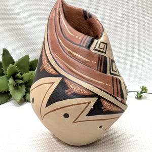 May include: A hand-painted, Native American-style pottery vase. The vase features a cream-colored base with geometric designs in black, brown, and tan. The top portion has a unique, folded shape with additional painted patterns.