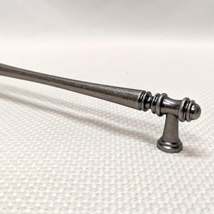 May include: A dark gray cabinet pull with a decorative design. The handle is a long, straight bar with rounded ends and a decorative center. The pull is approximately 6 inches long.