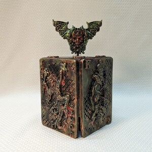 May include: A decorative box with a skull and bat design on top. The box is made of metal and has a distressed finish. The box is decorated with playing card symbols and the letter "K".