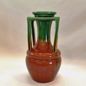 May include: A brown and green ceramic vase with a unique design. The vase has a wide base and a narrow neck with a green top. The vase has four handles that are attached to the neck and extend down to the base.