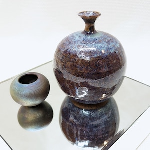 May include: A brown and blue ceramic vase with a round body and a narrow neck, next to a smaller brown and blue ceramic bowl. Both pieces are sitting on a mirrored surface.