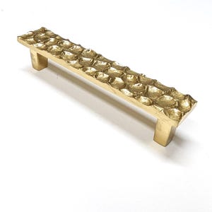 May include: A gold-colored cabinet pull with a textured, honeycomb-like design. The rectangular handle has two square posts for mounting. The hardware is made of metal and is suitable for furniture or cabinetry.